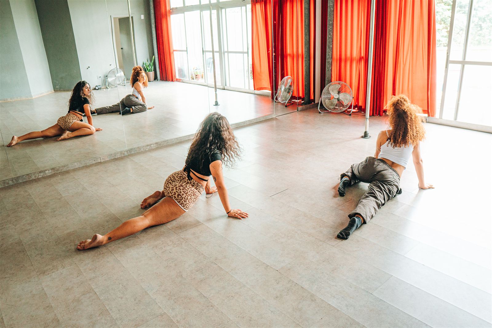 Camila e aluna alongando de frente para o espelho em estúdio com cortinas laranja