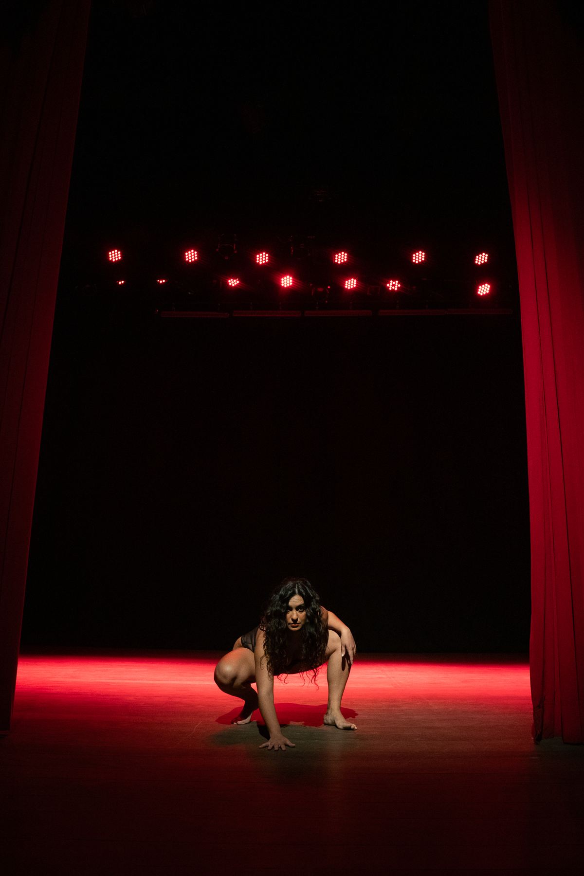 Camila agachada no palco sob varas de luz vermelha
